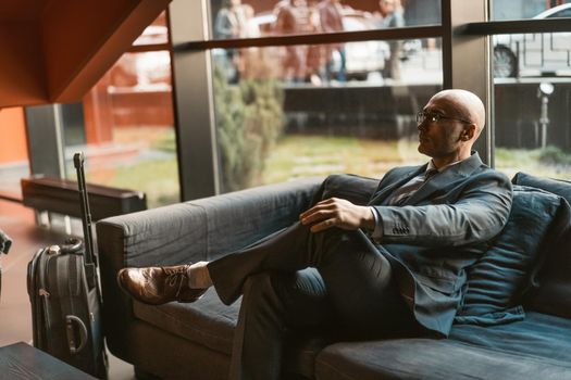 Waiting in hotel lobby respectable businessman looking away while waiting for check in or check out attending business forum. Handsome wearing business suit sitting in dark sofa in reception area.