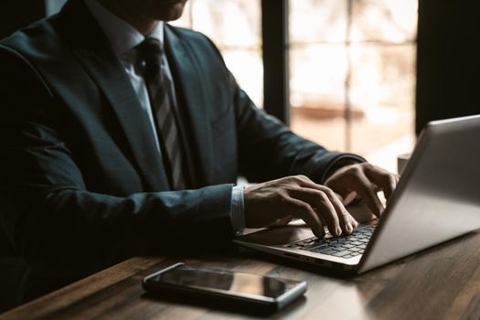 Close up. Businessman in expensive business suite working on laptop and smartphone next to it with windows on background in modern office. Man working on laptop in office.
