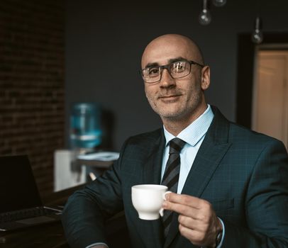 Mature business man in a business suit, holding a cup of coffee in a office building cafeteria having and looking at camera wearing expensive suit and eye glasses. Business concept.
