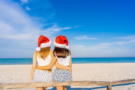 View from back of unrecognizable young couple of lover girls embracing each other sitting on beach wooden fence wearing Santa Claus hat in winter holidays vacations at ocean sea resort. Bright filter