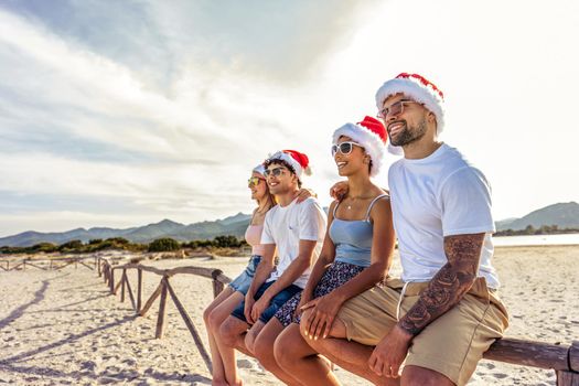 Perspective wide angle view of tattooed beautiful guy embracing his Hispanic girlfriends sitting on a wooden fence on a tropical beach enjoying holidays vacation sunglasses and Santa claus hat