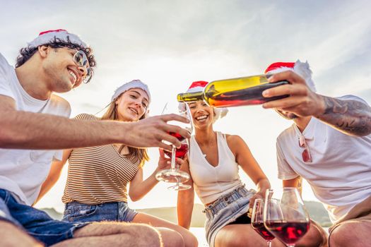 Tattooed guy pouring red wine from bottle wearing Santa Claus red hat at sunset on a wooden pier in ocean sea resort for winter holidays vacations. Outdoor friends party