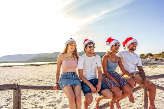 Happy people enjoying outdoor life in exotic vacation resort sitting on wooden fence on a white sand beach at sunset. Group of young friends having fun together Santa Claus hat at sea vacation resort