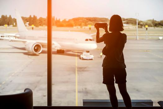 A young girl takes a photo on her phone before boarding the plane, the traveler goes on a trip, a silhouette at the airport by the window against the background of the aircraft.