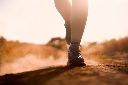 Female athletic legs in sneakers at sunset during outdoor jogging at sunset. A young girl warms up, she is engaged in trail running, view on her legs and clouds of dust close-up.