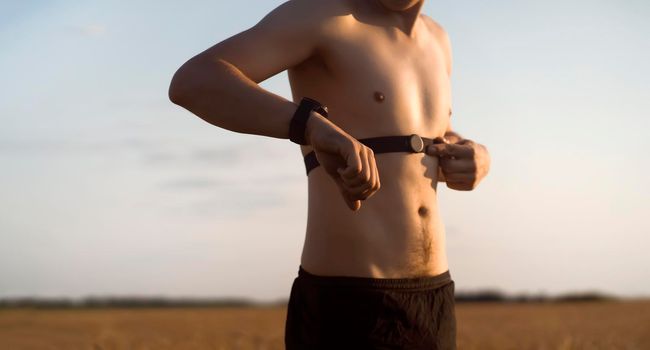 A young male athlete puts on a heart rate monitor and smart watch before jogging in nature. A man monitors his pulse and is engaged in a healthy running at sunset.