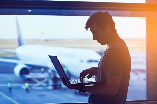 A young man is working on a laptop at the airport while waiting to board the plane. A man is engaged in business, buys tickets, studies and communicates via the Internet at sunset.