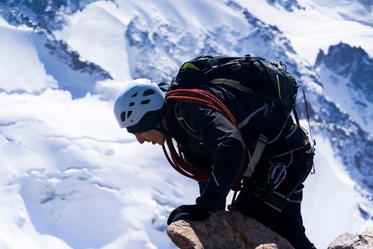 A young man traveler is engaged in mountaineering. Hiker in a helmet, with a rope climbs to the top, against the backdrop of a stunning view with snow-capped mountains.