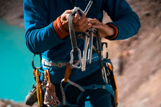 A man with climbing equipment holds an ice screw in his hands against the backdrop of a mountain lake and rocks.