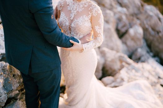 Groom hugs bride by the waist while standing on the stones. Close-up. High quality photo