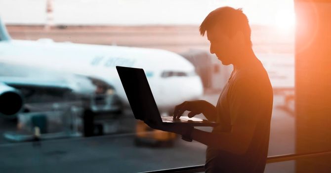 A young man is working on a laptop at the airport while waiting to board the plane. A man is engaged in business, buys tickets, studies and communicates via the Internet at sunset.