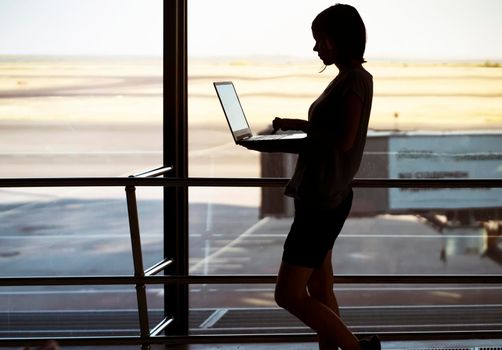 Silhouette of a young girl with a laptop standing near the window at the airport, a woman works online, studies, is engaged in a business project remotely during her flight.