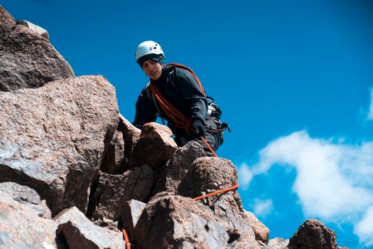 A young man traveler is engaged in mountaineering. Hiker in a helmet, with a rope climbs to the top, against the backdrop of a blue sky.