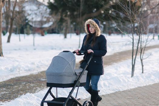 Beautiful mother walking in the park with her little baby in stoller. Woman dressed in blue jaket with hood and jeans. Warm boots.