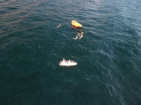 Happy people swim on air mattress behind a high speed water bike. Tourists ride on inflatable watercraft boat. Sea attraction. Water boat skating people on large Inflatable raft. Aerial copter view