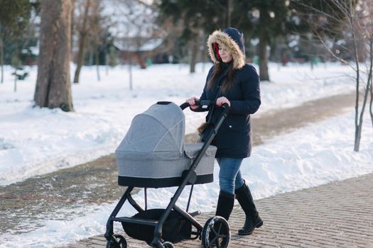 Beautiful mother walking in the park with her little baby in stoller. Woman dressed in blue jaket with hood and jeans. Warm boots.