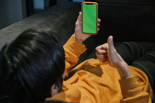 An Asian guy lays on sofa in a small office and communicates via video link through a mobile phone with green screen. The concept of small business and online communication.