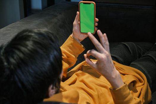 An Asian guy lays on sofa in a small office and communicates via video link through a mobile phone with green screen. The concept of small business and online communication.