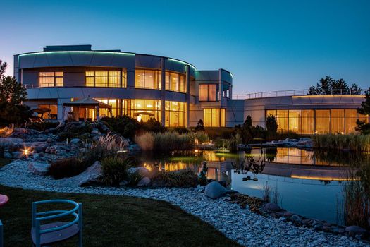 Twilight view of modern stylish two-story country house with lighted wide windows and well-groomed adjacent territory with small artificial pond in summer