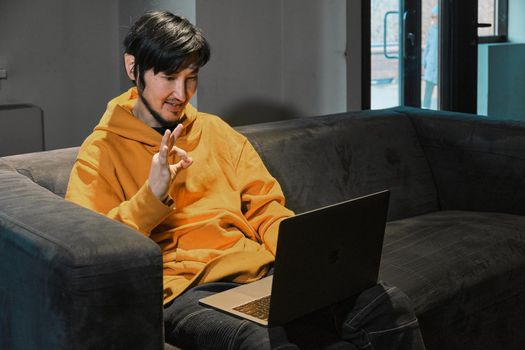 An Asian guy sits on sofa in a small office and communicates via video link through a laptop. The concept of small business and online communication.