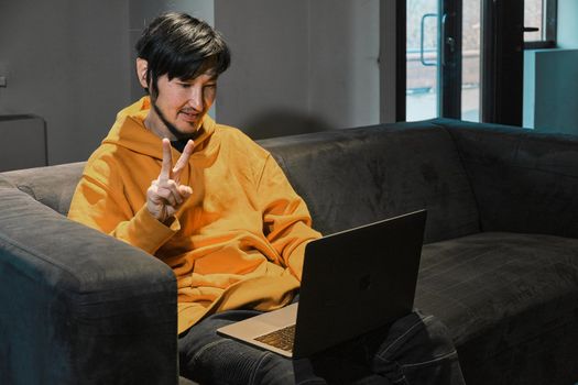 An Asian guy sits on sofa in a small office and communicates via video link through a laptop. The concept of small business and online communication.