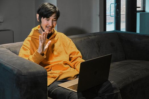 An Asian guy sits on sofa in a small office and communicates via video link through a laptop. The concept of small business and online communication.