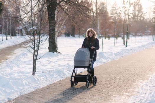 Beautiful mother walking in the park with her little baby in stoller. Woman dressed in blue jaket with hood and jeans. Warm boots.