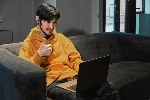 An Asian guy sits on sofa in a small office and communicates via video link through a laptop. The concept of small business and online communication.