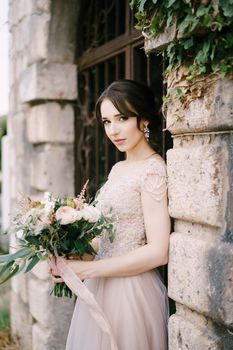 Bride with bouquet of flowers stands at the forged gate in a stone fence. High quality photo