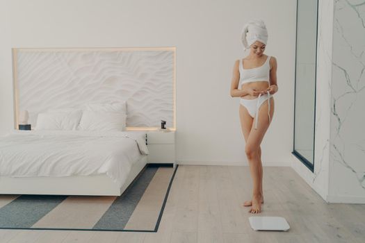 Beautiful slim fit female model posing in white underwear with measuring tape in hand next to electronic scales on floor in stylish bedroom background, full length. Weight loss and dieting concept