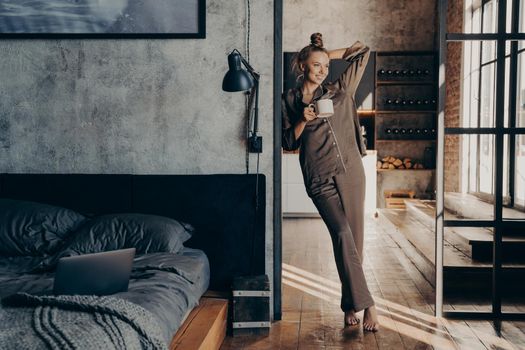 Young pretty happy woman wearing brown satin pajama, holding coffee cup mug while standing in bedroom doorway after waking up in morning at home, female starting new day