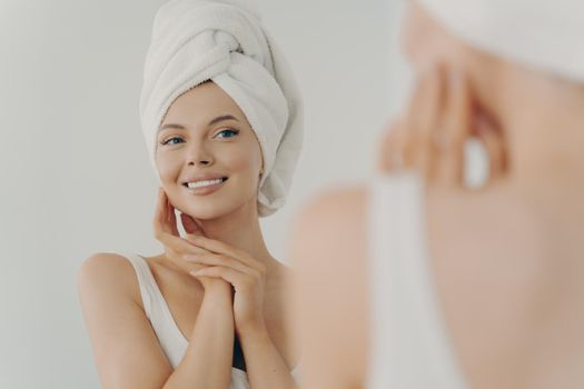 Beautiful young woman after shower looks in mirror gently touching soft healthy glowing face skin, happy millennial female applying beauty products during daily facial procedures, skincare concept