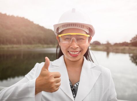 Female engineer in a helmet looks and smiles at the camera while holding a digital tablet with her thumb up at a dam construction site to generate electricity.