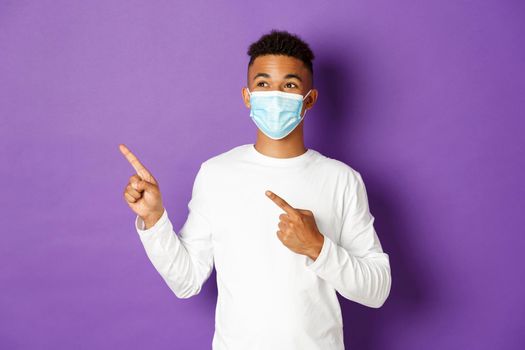 Concept of coronavirus, quarantine and lifestyle. Handsome young african-american man in medical mask, pointing and looking at upper left corner, showing advertisement, posing over purple background.