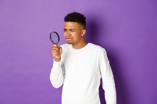 Image of young african-american male student looking for something with magnifying glass, squinting as cant see something small, standing over purple background.