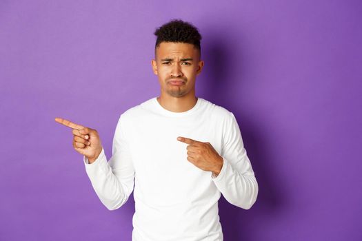 Portrait of sad and disappointed african-american young man, complaining and sulking, pointing fingers left at something bad, standing over purple background.