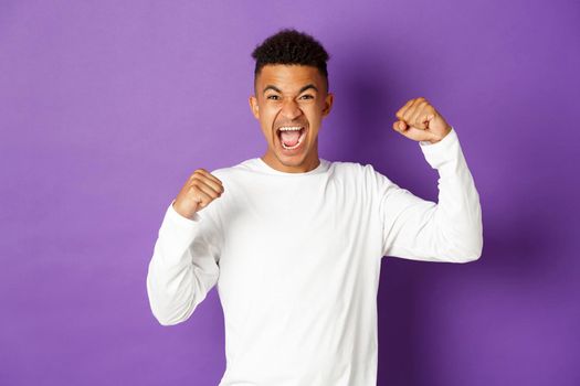 Image of happy african-american guy, rooting for sports team, raising hands up and shouting for joy, say yes and celebrating victory, triumphing over purple background.