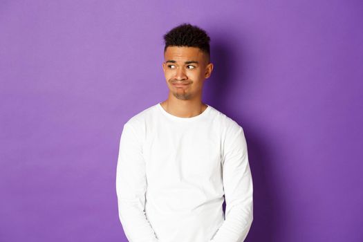 Image of disappointed and sad african-american man, looking left and grimacing, standing over purple background.
