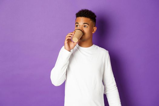 Portrait of young modern african-american guy drinking coffee, looking at upper left corner, standing over purple background.