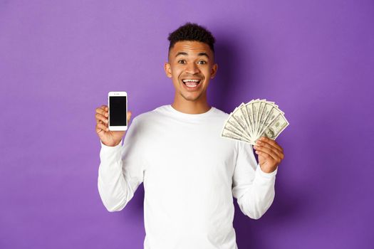 Portrait of cheerful african-american man, showing mobile phone screen and money, standing against purple background.