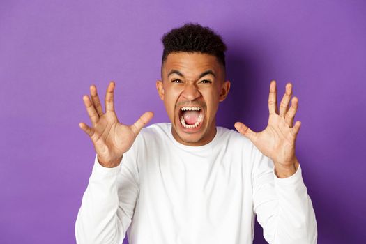Image of pissed-off african american guy yelling at someone, raising hands and looking with hatred and anger, standing over purple background.