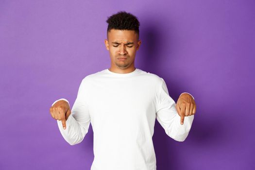 Portrait of skeptical and disappointed african-american male model in white sweatshirt, frowning and pouting upset, pointing and looking down, standing over purple background.