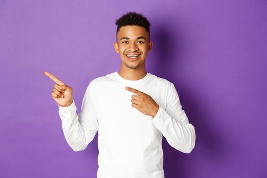Cheerful african-american man in white sweatshirt showing copy space for logo, pointing fingers left, smiling at camera, standing over purple background.