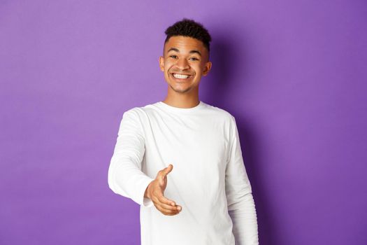 Image of handsome young african-american man, smiling friendly while extending hand for handshake, greeting someone, standing over purple background.