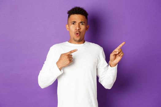Portrait of confused and shocked african-american guy in white sweatshirt, pointing fingers right and frowning, showing something strange, standing over purple background.