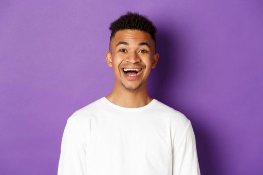 Close-up of joyful african-american man in white sweatshirt, laughing and looking amazed at camera, standing over purple background.