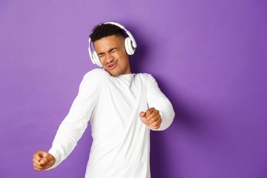 Image of carefree african-american guy dancing, listening music in wireless headphones, standing over purple background.