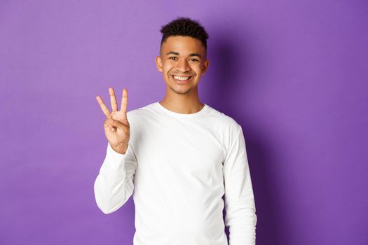 Image of cheerful african-american guy in white sweatshirt, showing three fingers and smiling, standing over purple background.