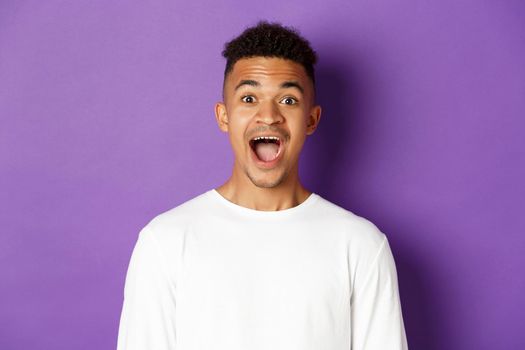 Close-up of amazed african-american young man, open mouth and saying wow, looking fascinated at camera, standing over purple background with copy space.