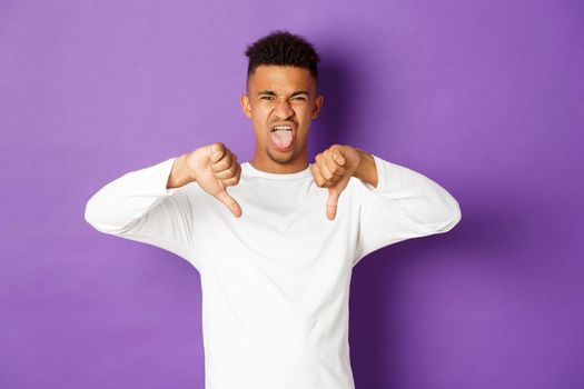 Image of skeptical and disappointed african-american young man, express disgust, showing tongue and thumb-down from aversion and dislike, standing over purple background.